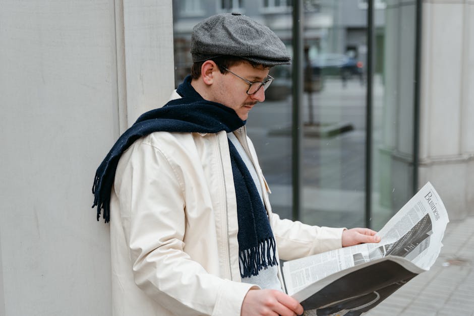 A man in a flat cap and scarf reads a business newspaper outdoors on a chilly day.