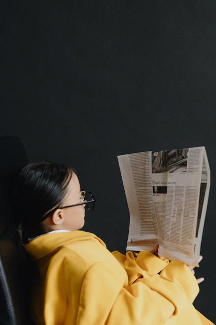 A young girl with eyeglasses reads a newspaper indoors, wrapped in a yellow coat.