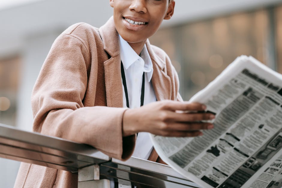 Crop happy African American businesswoman in formal clothes reading newspaper and looking away with smile while leaning on railing outside blurred modern bui…